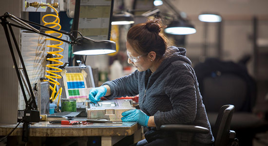 A person wearing safety glasses and gloves, working at a lit workstation with tools and materials, in a workshop setting.